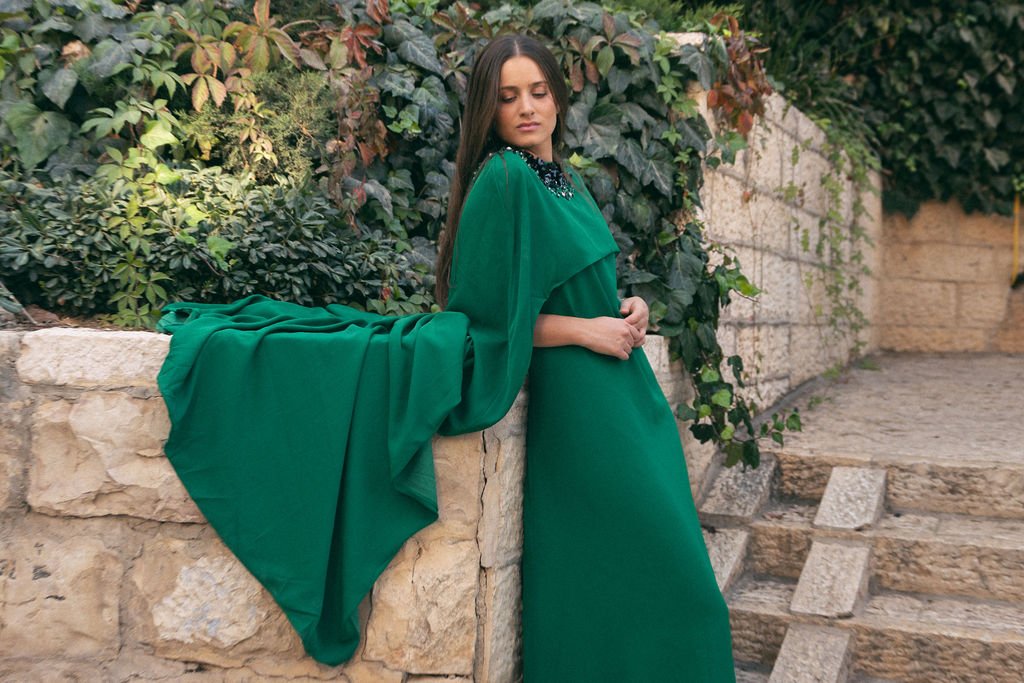 A woman in a long green dress with puffy sleeves standing next to a stone wall with greenery in the background.