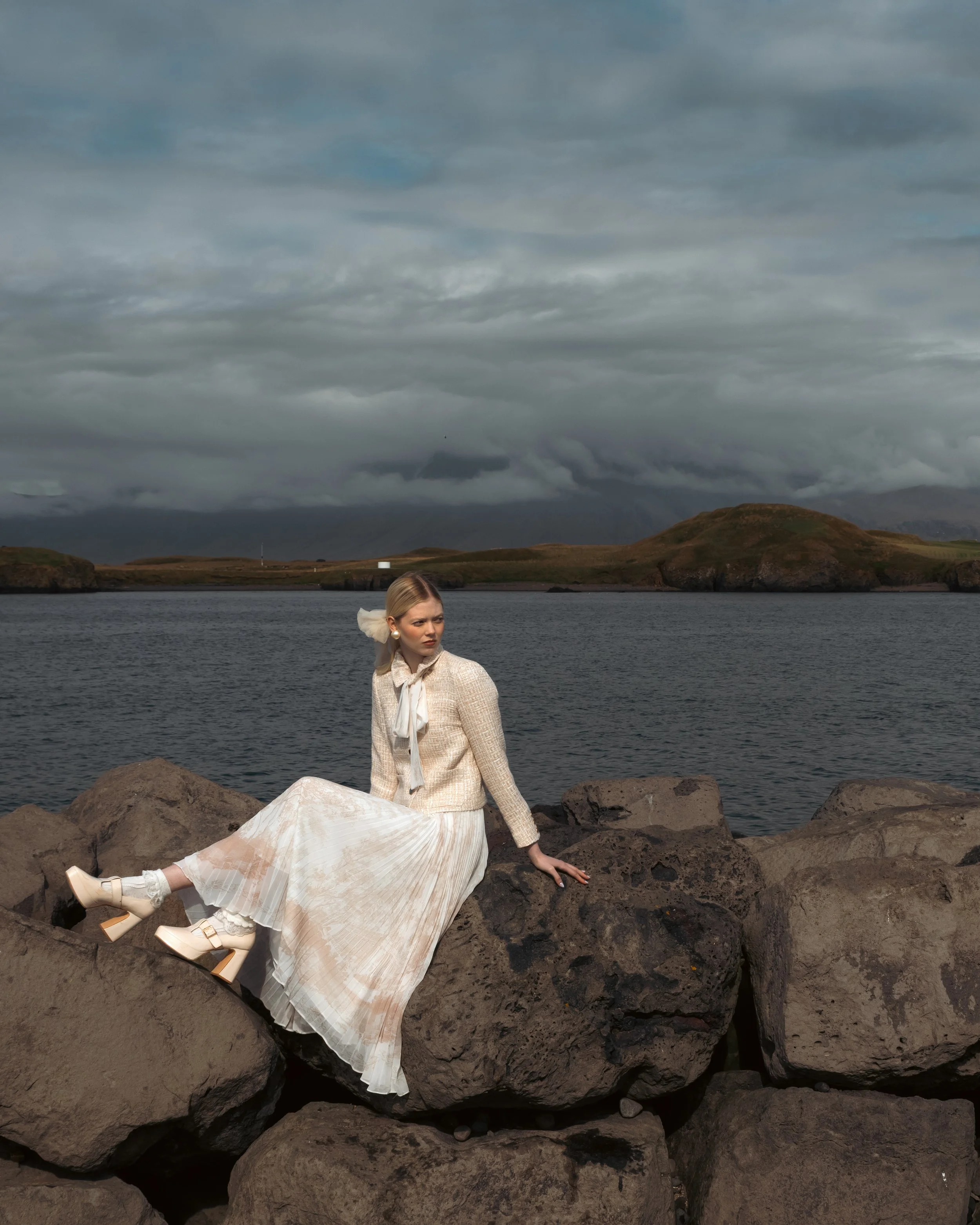 A woman in a vintage white dress and cream-colored jacket sits on rocks beside a body of water, with hills and cloudy sky in the background.