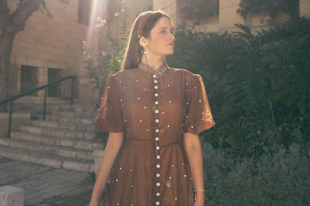 A woman standing outdoors at sunset, wearing a brown dress with sheer puffed sleeves and pearl embellishments, with a staircase and greenery in the background.