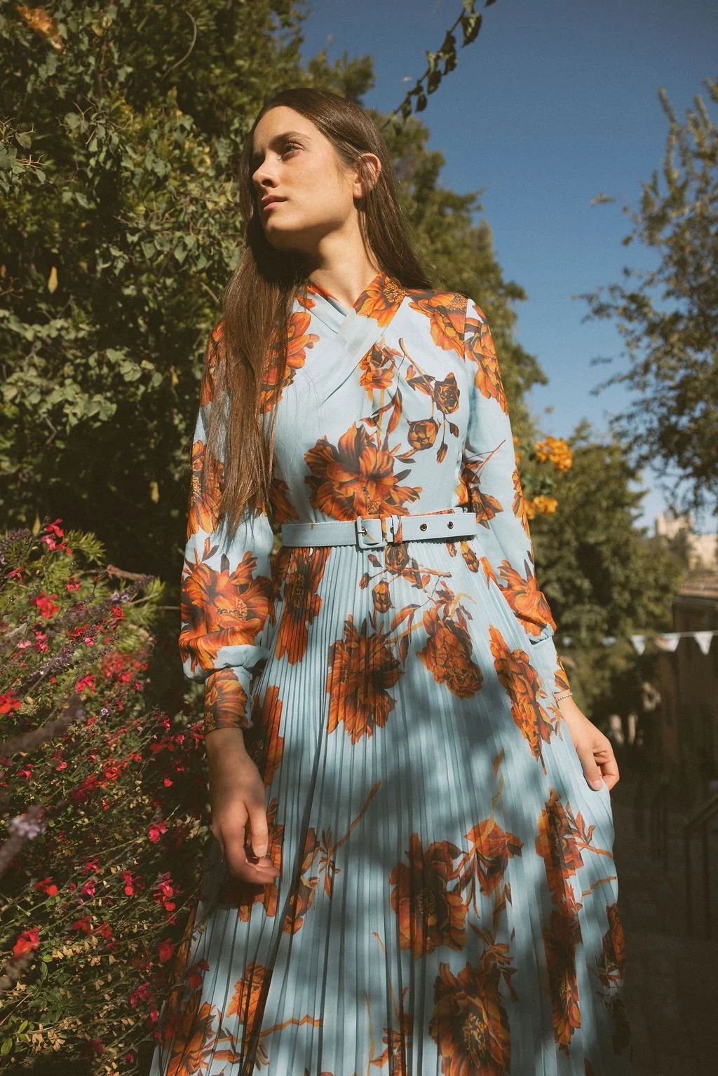 A woman with long brown hair stands outdoors among trees and bushes, wearing a light blue dress with orange floral patterns and a matching belt. The sky is clear and blue.