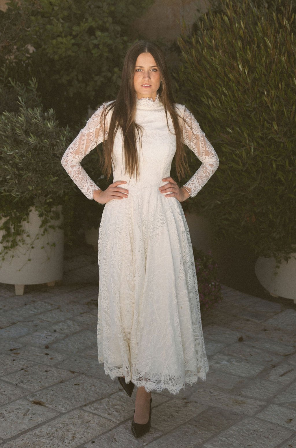 A woman standing outdoors on a stone pathway, wearing a long white lace dress and black heels, with her hands on her hips and surrounded by greenery.