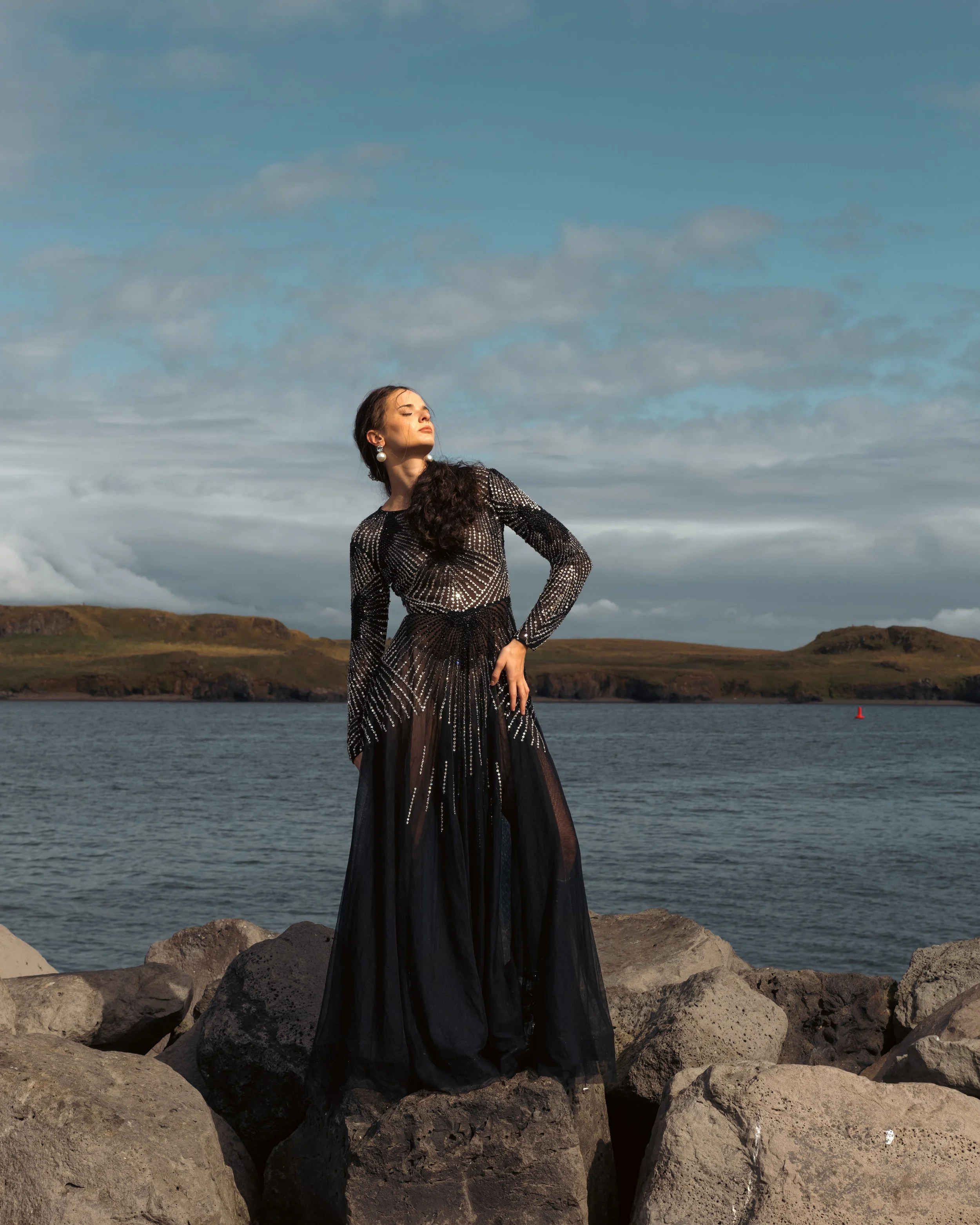 A woman in an elegant black dress with sparkly embellishments standing on rocks by a body of water, with rolling hills and cloudy sky in the background.