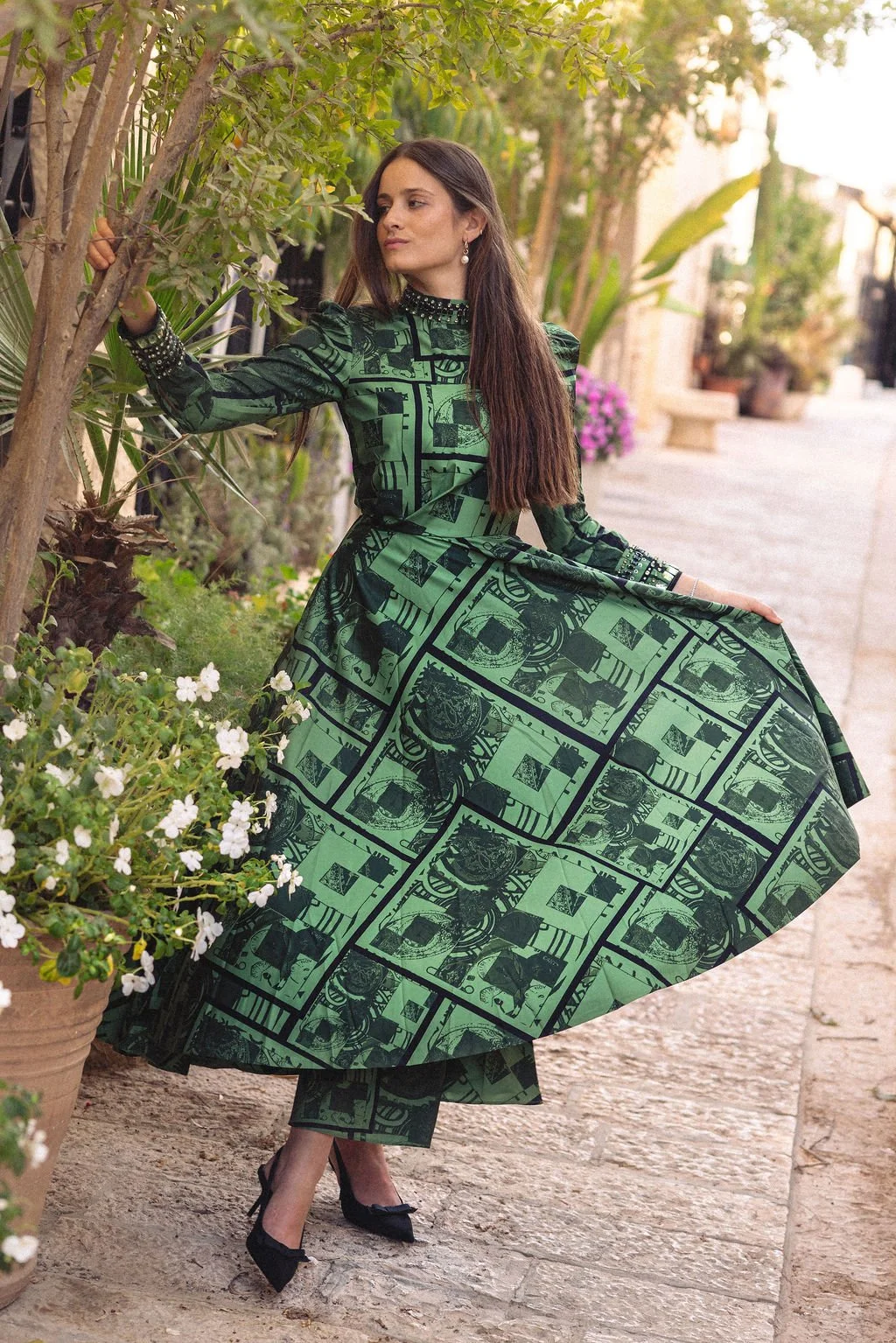 A woman in a green and black printed dress with a high collar, standing outdoors next to a tree, holding the edge of her dress and posing on a pathway lined with plants and flowers.