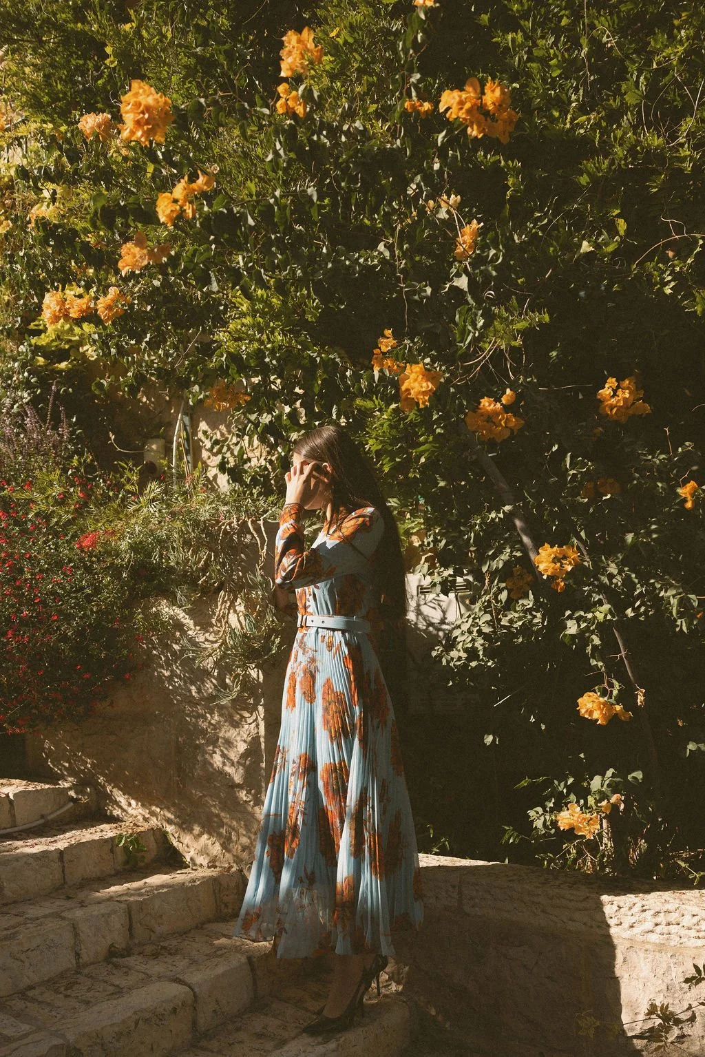 A woman in a floral dress standing on stone steps, surrounded by lush green foliage and yellow flowers, with sunlight casting shadows.