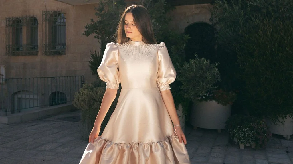 A woman standing outdoors in a satin dress with puff sleeves and a ruffled hem, with trees and buildings in the background during daylight.