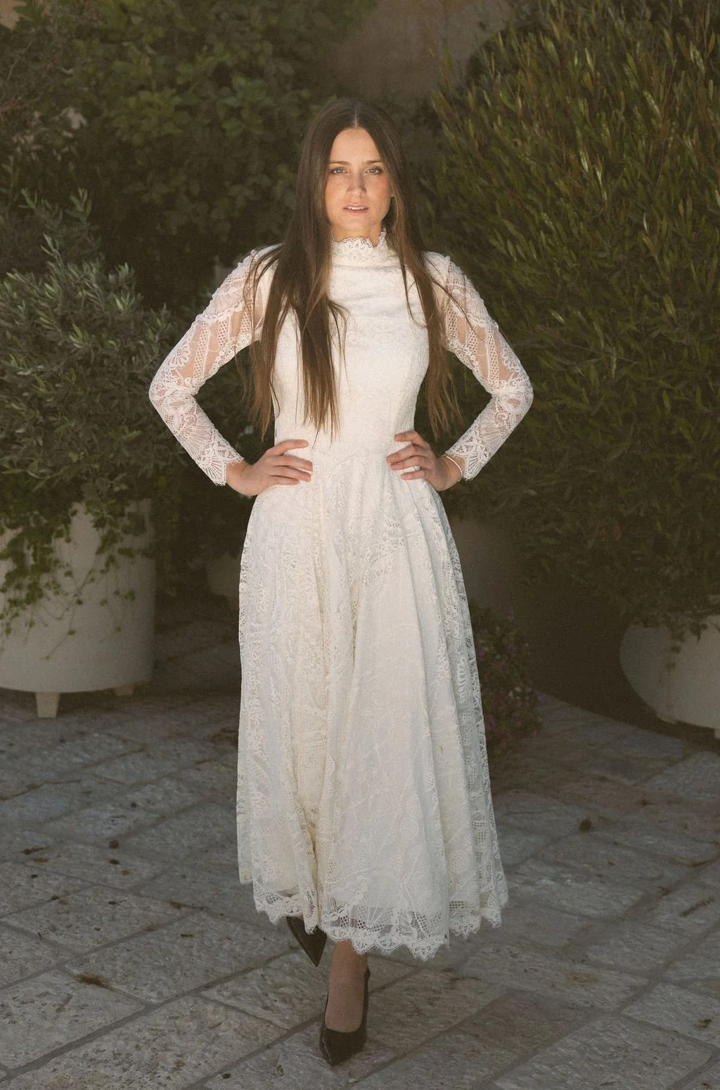 A woman with long brown hair wearing a white lace dress with long sleeves, standing outdoors on a stone pathway with greenery in the background.