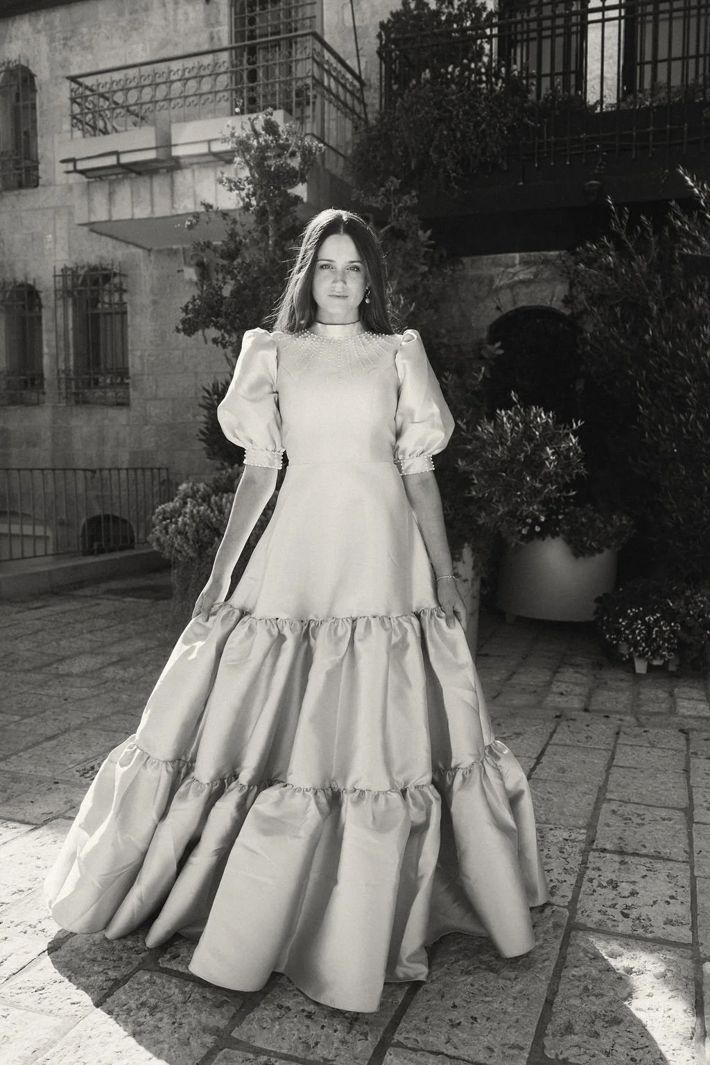 A woman in a long, elegant, vintage-style gown with puffed sleeves, standing outdoors on a cobblestone street at night, surrounded by potted plants and old buildings.