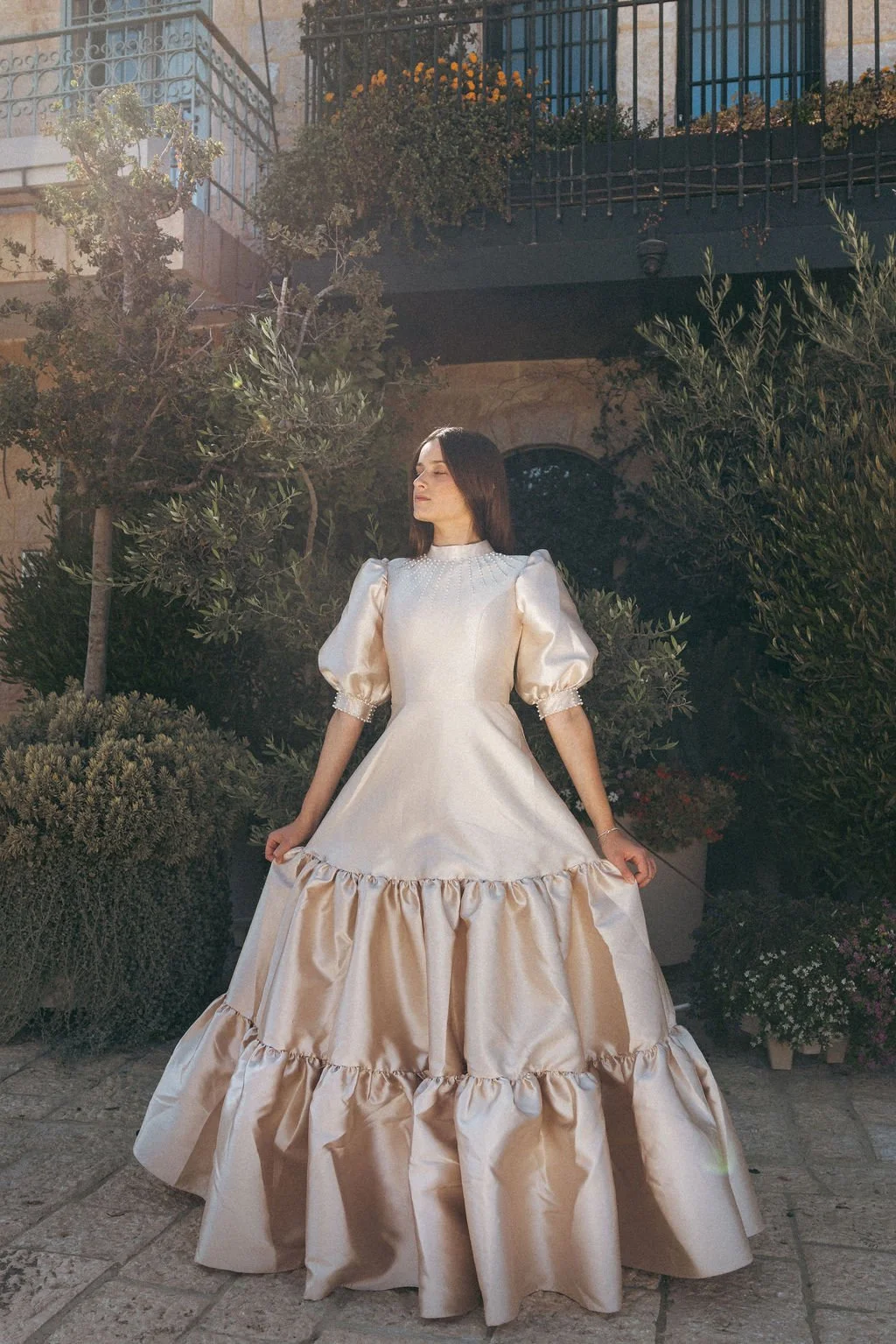 A woman in a vintage style satin wedding gown with puffed sleeves and ruffle details stands outdoors in front of lush greenery, with a building and balcony in the background, illuminated by soft sunlight.