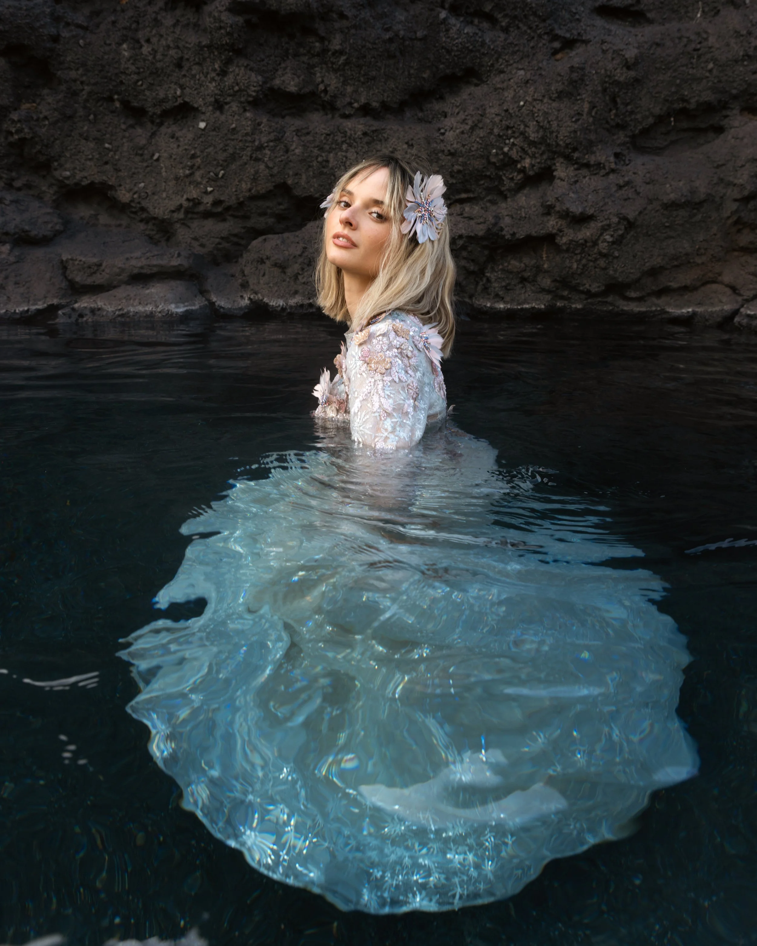 A woman with blonde hair is partially submerged in dark water, near a rocky wall, wearing a floral dress and accessorized with a large flower hairpiece.