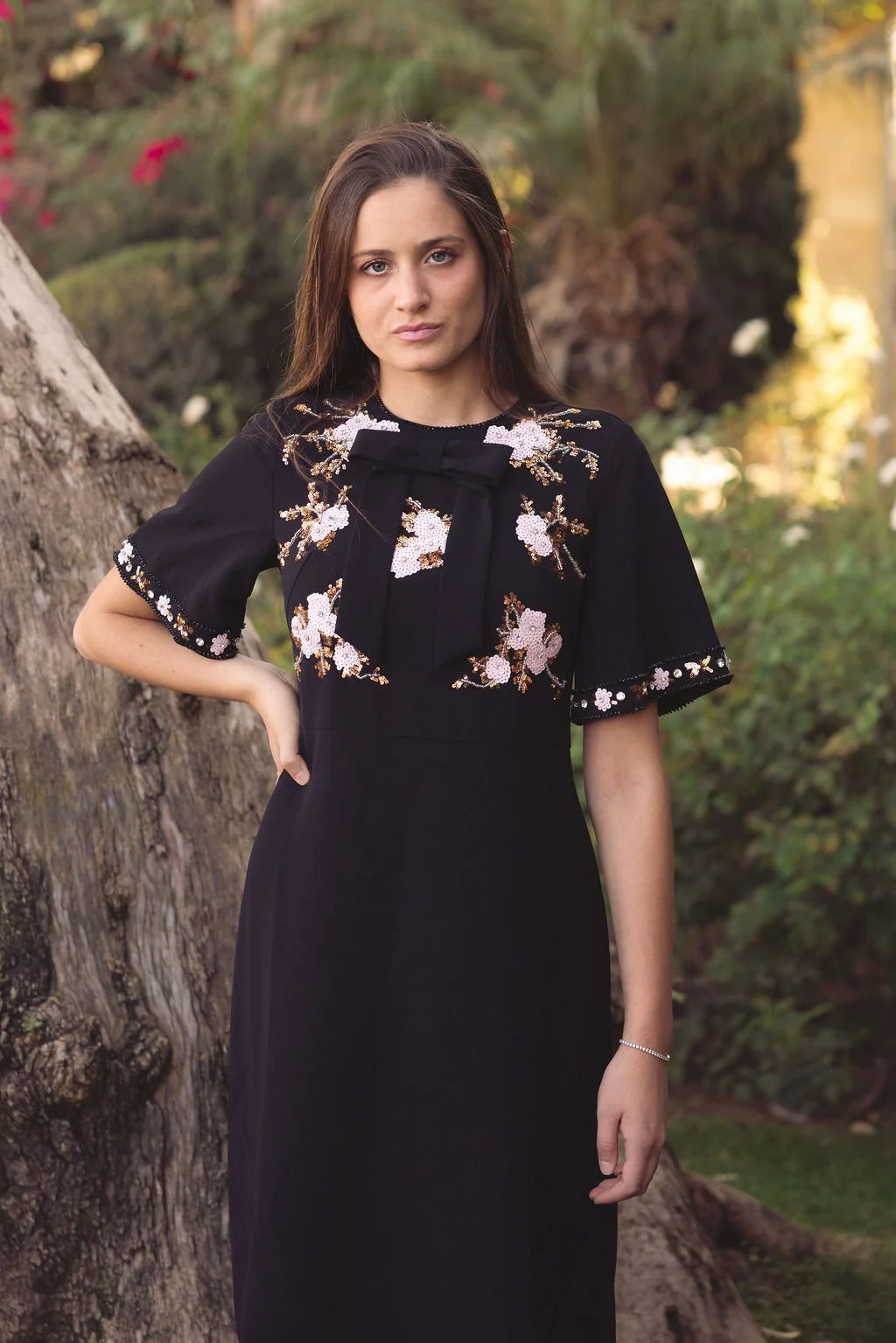 A young woman with long brown hair and light skin wearing a black dress with embroidered white and pink floral patterns, standing outdoors near a large tree with a blurred green and yellow background.