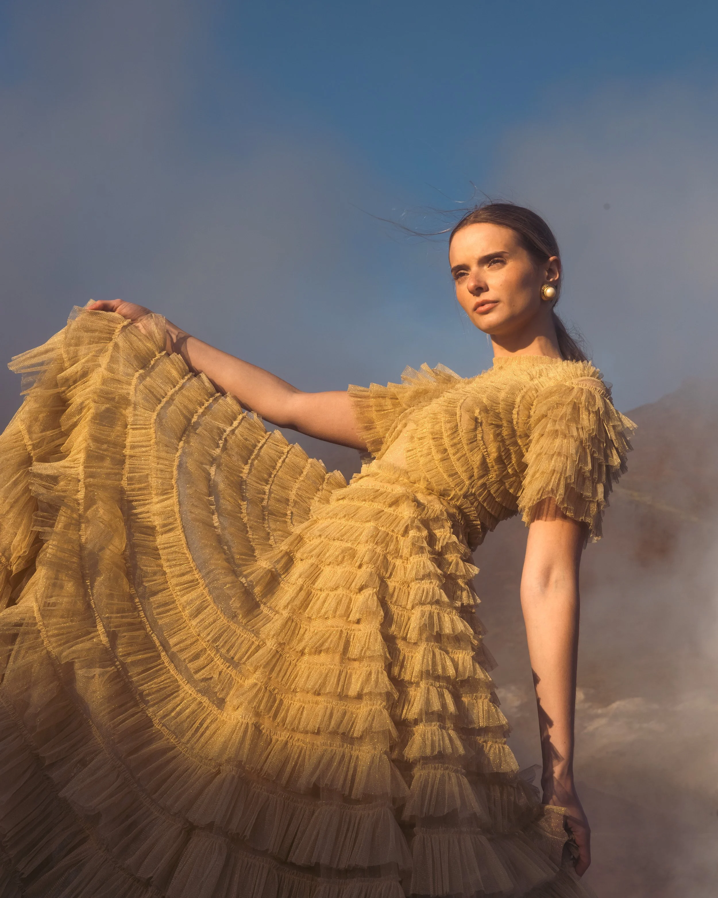 A woman in a yellow, ruffled, flowing dress stands outdoors against a cloudy sky, gazing into the distance.