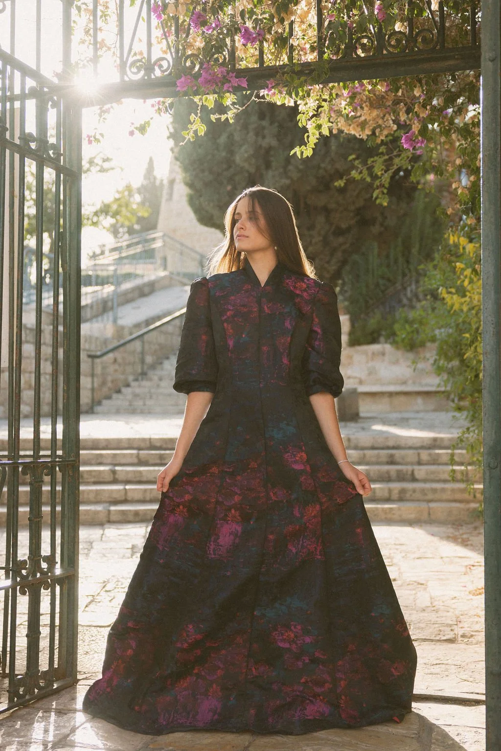 A woman in a dark, floral-patterned dress standing in a doorway with sunlight shining behind her.