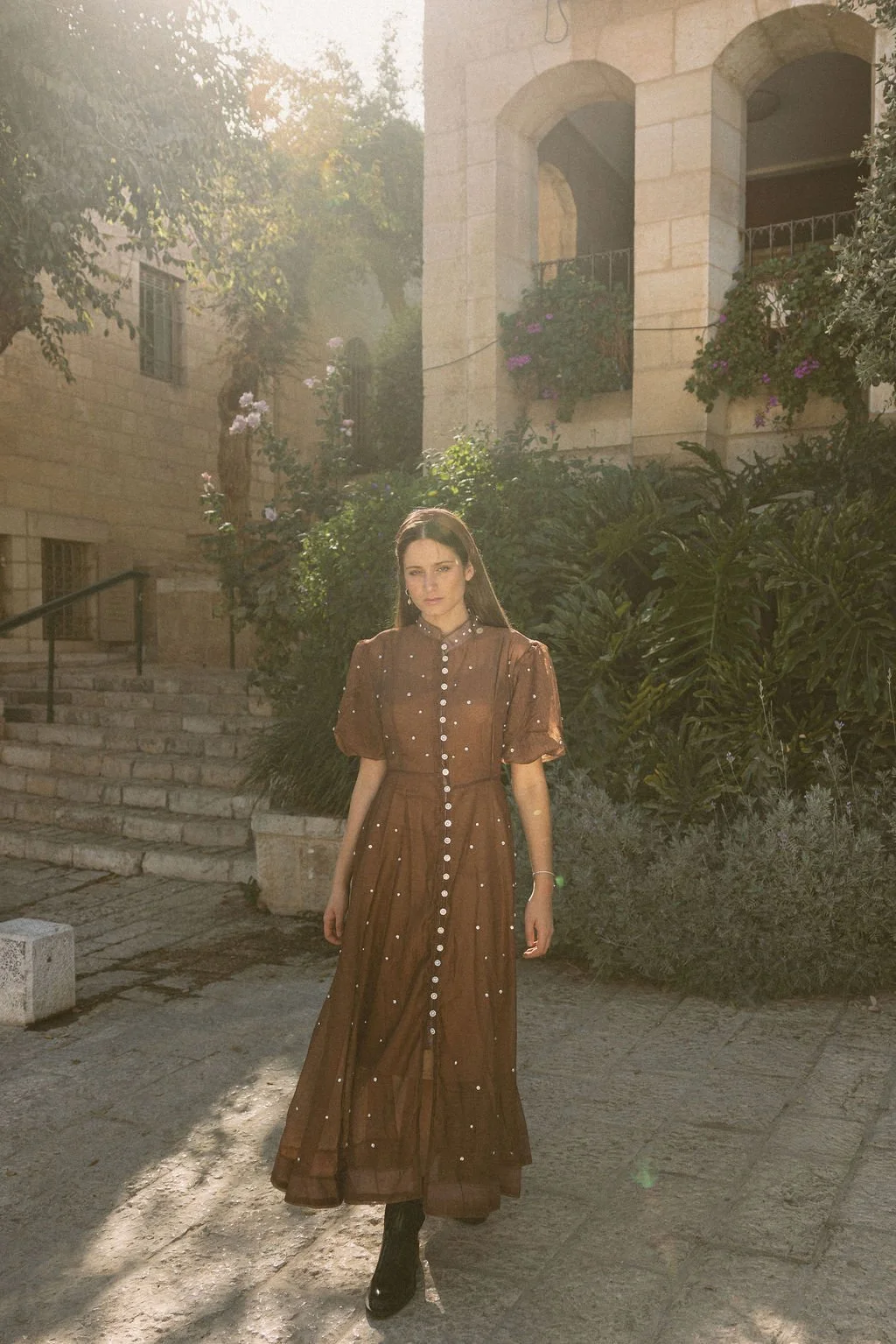 A woman in a long, brown, button-down dress with puffed sleeves stands on stone steps amidst greenery, with sunlight filtering through trees behind her.