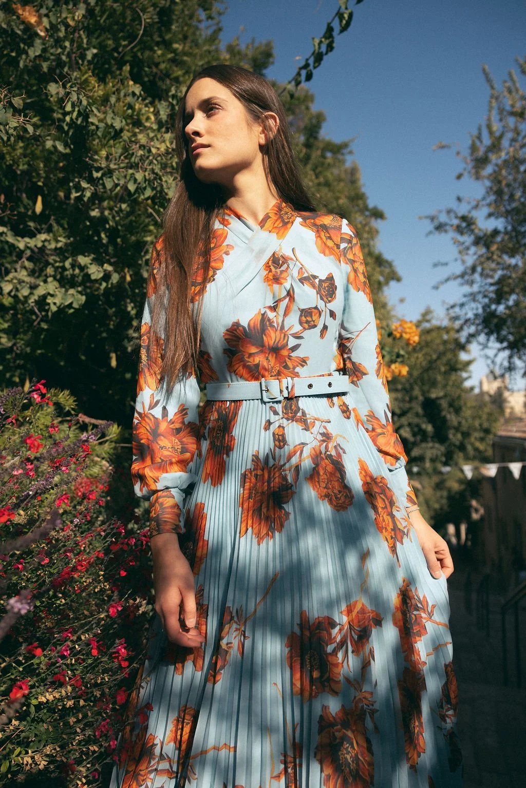 Woman with long brown hair in a blue floral dress standing outdoors among greenery and flowers on a sunny day.