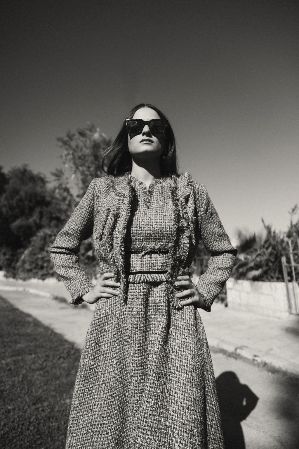 A woman wearing a textured coat and oversized sunglasses standing outdoors with hands on hips, in a black and white photograph.