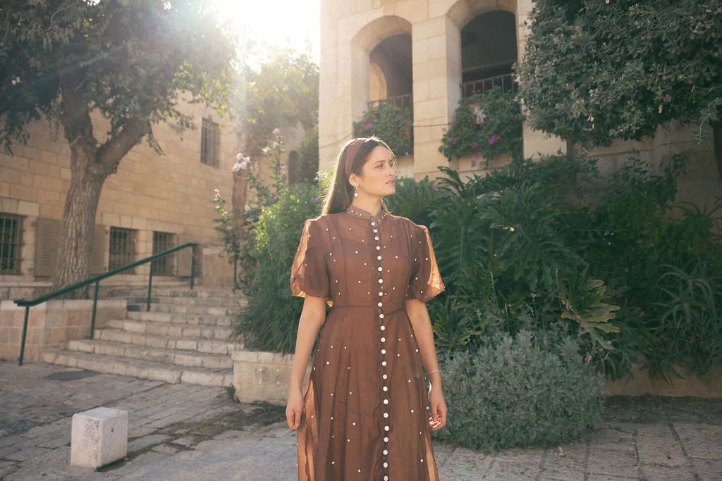 A woman in a brown dress with pearl accents standing outdoors in a historical stone building area with stairs and greenery, illuminated by sunlight.