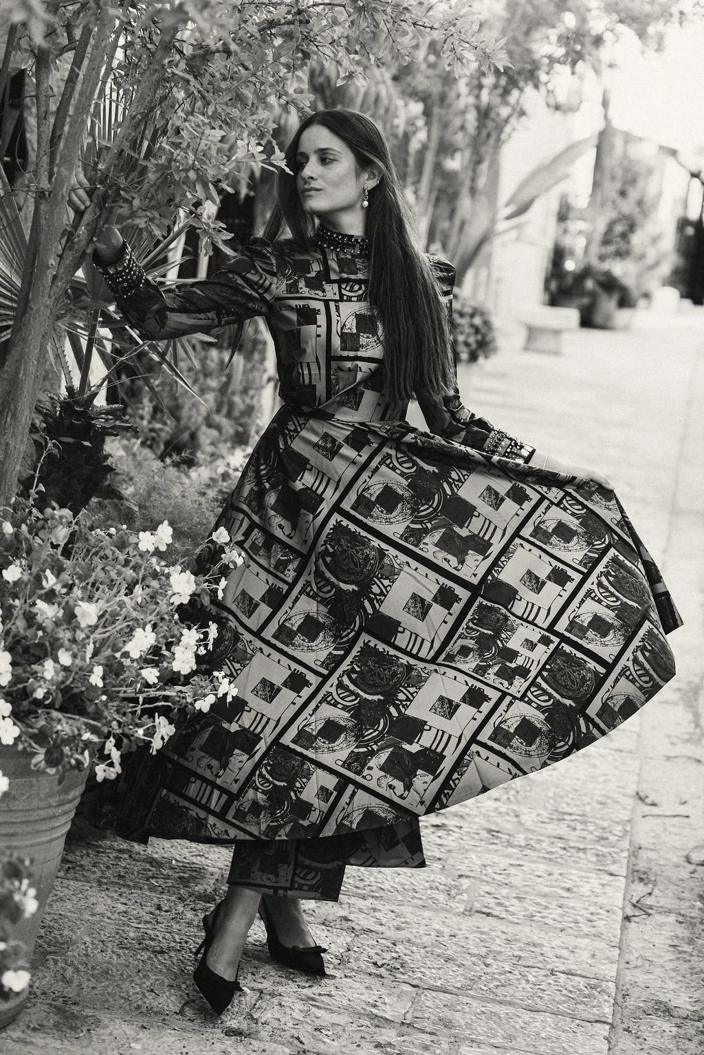 A woman in a patterned dress standing outdoors near some plants and flowers, holding a branch, in a garden or park setting, black and white photo