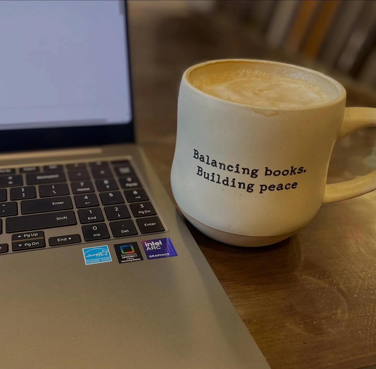 A coffee mug with the words 'Balancing books. Building peace' written on it, placed next to a laptop on a wooden table.