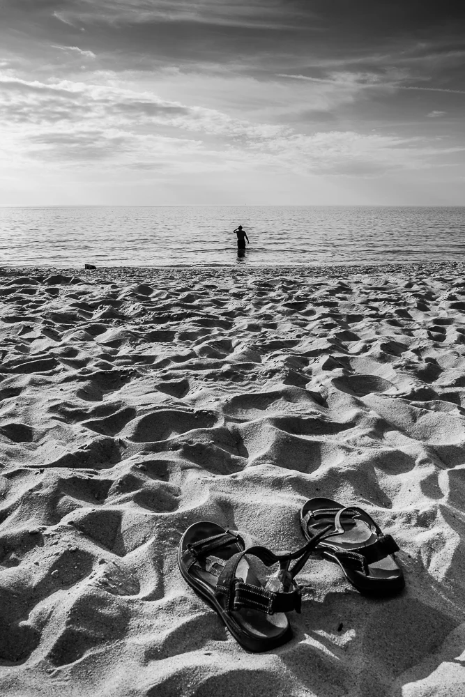 candid photo of sandals on beach and man walking into water