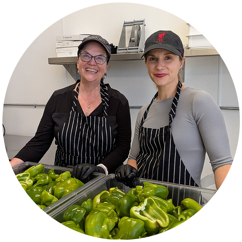 Two people volunteering in the kitchen in aprons standing by chopped green peppers