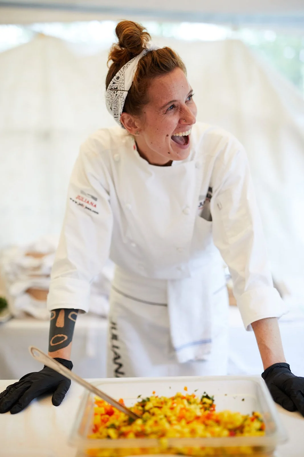 Juliana Trujillo Escobar  in a chef's uniform, smiling and laughing, standing behind a table with a dish of colorful food, wearing a bandana and black gloves.