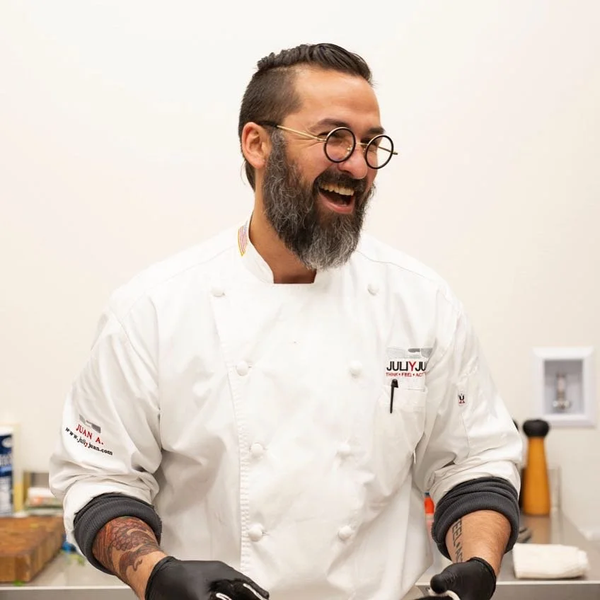 Juan Andres Rodriguez  smiling  with glasses and a beard wearing a white chef coat, standing in a kitchen.