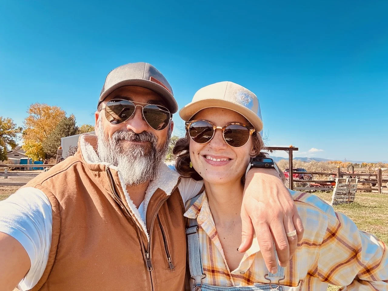 Juli y Juan in a casual picture in Colorado.  outdoors taking a selfie on a sunny day, both wearing sunglasses and hats, with trees, a fence, and parked cars in the background.
