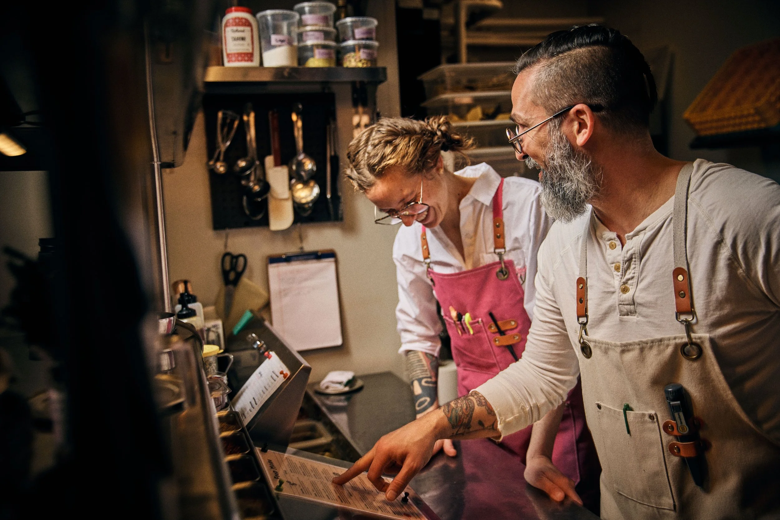 Two chefs, a woman and a man, are laughing and looking at a menu or screen in a kitchen. They are wearing beige and pink aprons and appear to be happy. The background shows kitchen utensils and shelves.