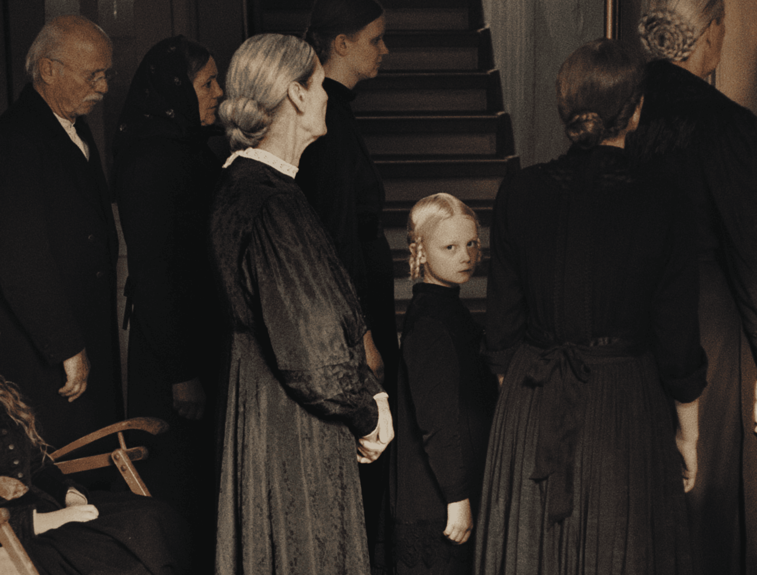 A group of people standing in a line indoors, dressed in dark clothing. The scene appears to be somber and reflective, possibly at a funeral or memorial service.