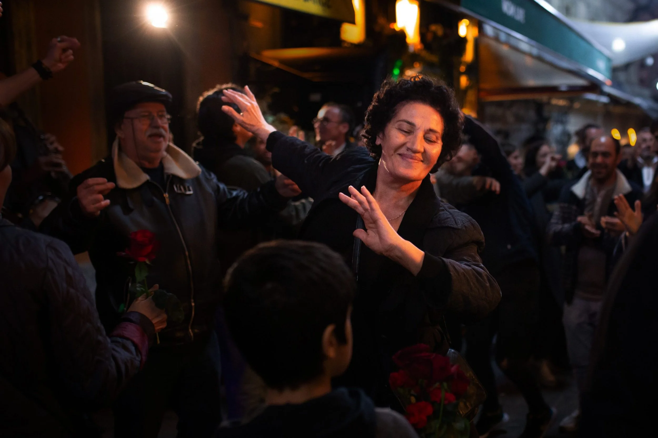 A film still from Crossing. A woman is smiling, dancing, surrounded by a crowd at night. he background shows warm ambient lighting.