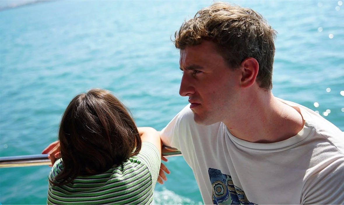 A man and a woman on a boat, with the woman leaning on the railing and the man looking at her, against a background of blue ocean water.