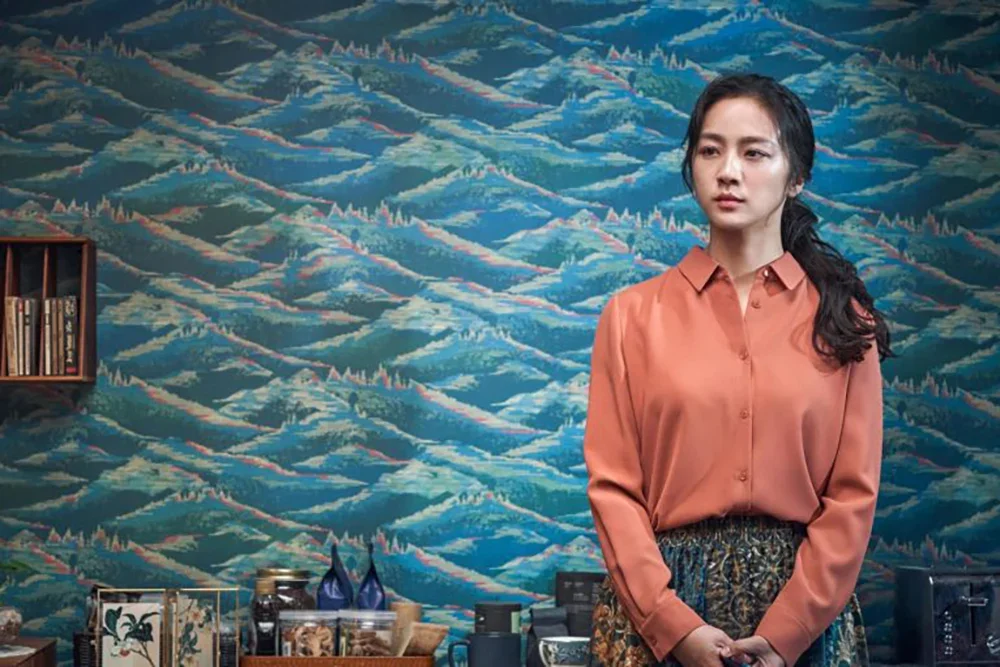 Still from Decision to Leave. A woman in a coral blouse and patterned skirt standing in front of a vibrant, abstract blue mountain landscape mural, with a shelf of books and decorative items behind her.