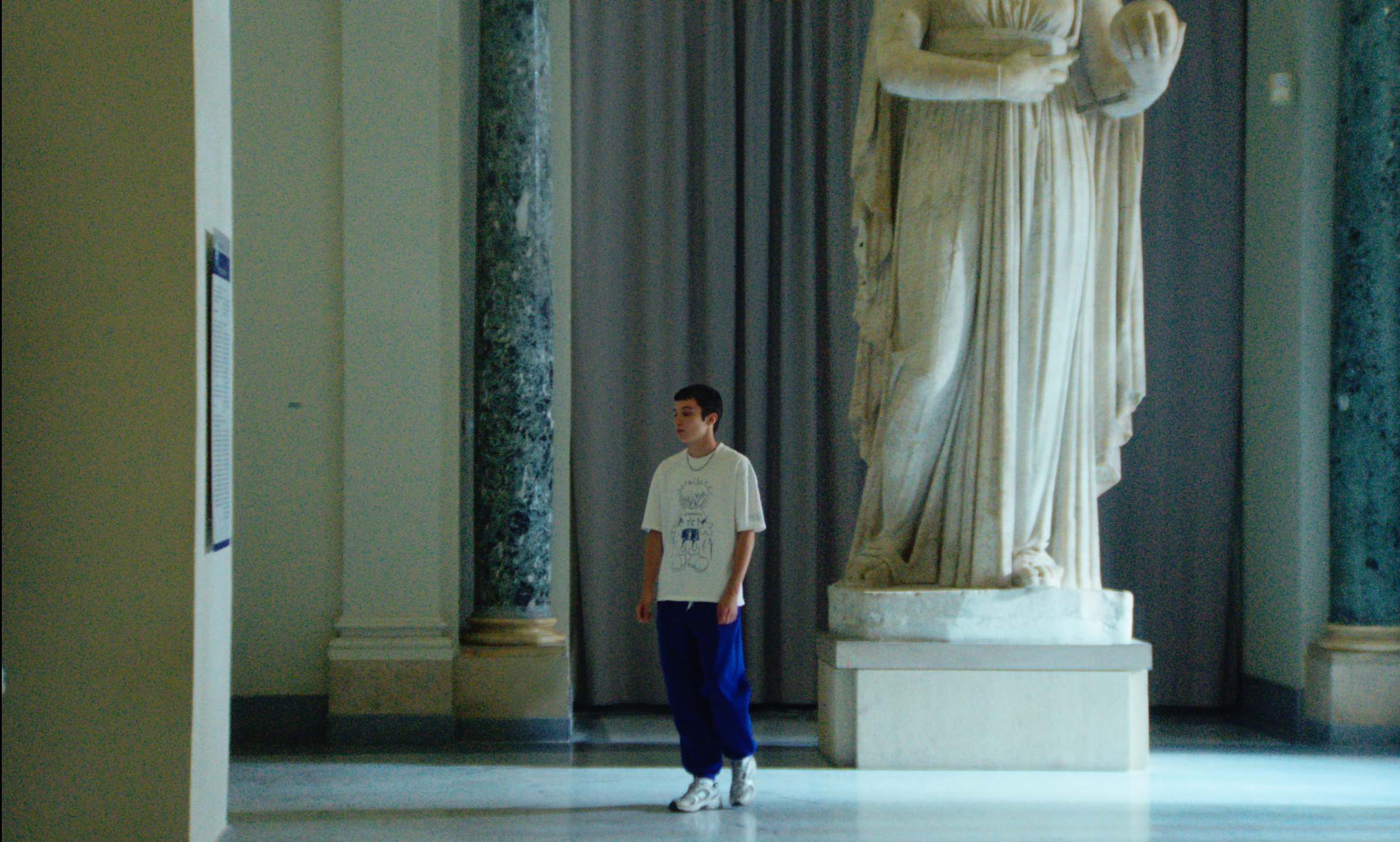 A film still from La Gradiva. A young boy walking inside a museum with classical sculptures and columns visible in the background.