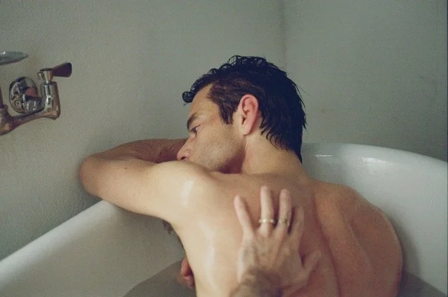 A film still from The Man I Love. A man with dark, wet hair is in a bath leaning against the edge, resting his head on his arm. Another man's hand is reaching from behind the camera to touch his back.