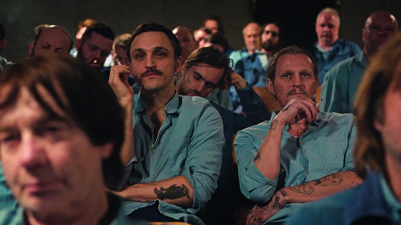 A group of men attending a meeting or seminar, sitting closely together, some holding cigarettes, with serious and attentive expressions, while one man is in focus with a mustache, sitting with his hand on his face, and another man with tattoos and a cigarette in his hand.