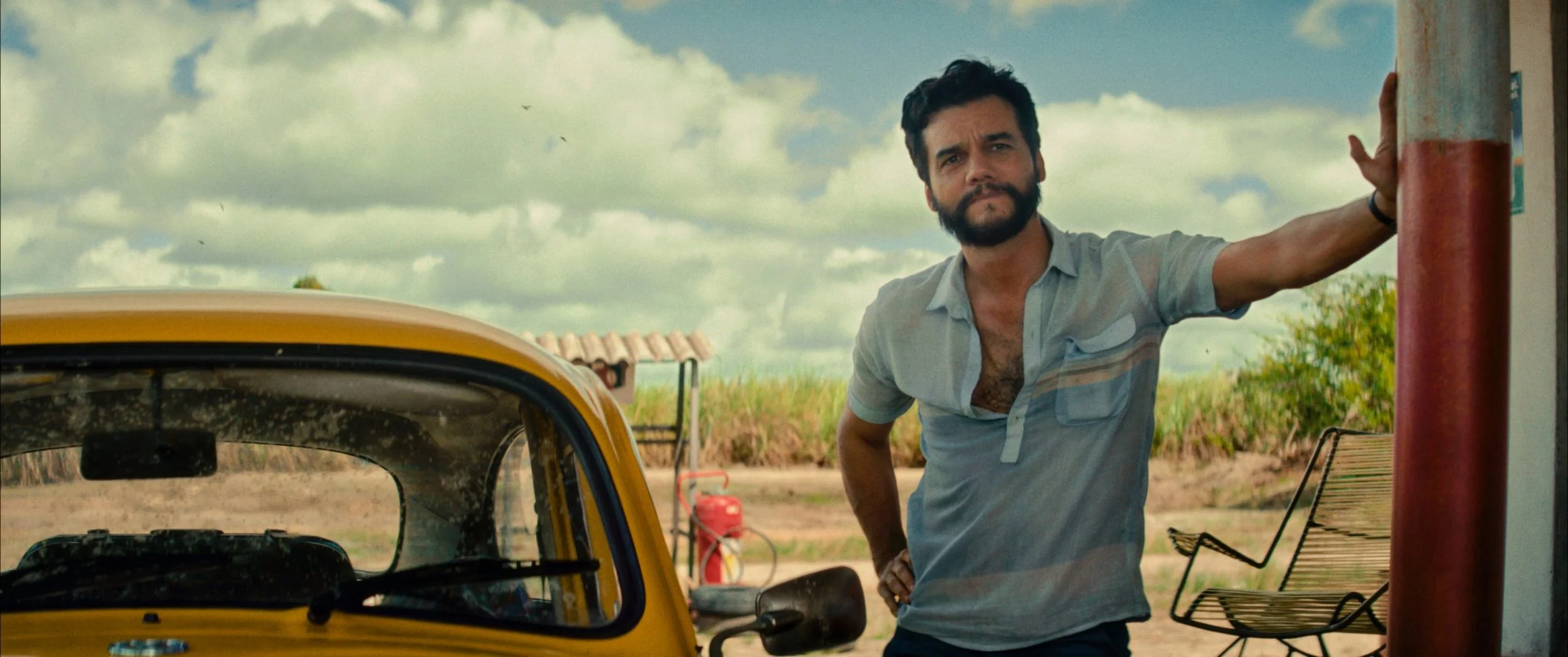 A man with dark hair and a beard standing outdoors next to a yellow vehicle, with his hand on a red pole, under a cloudy sky.