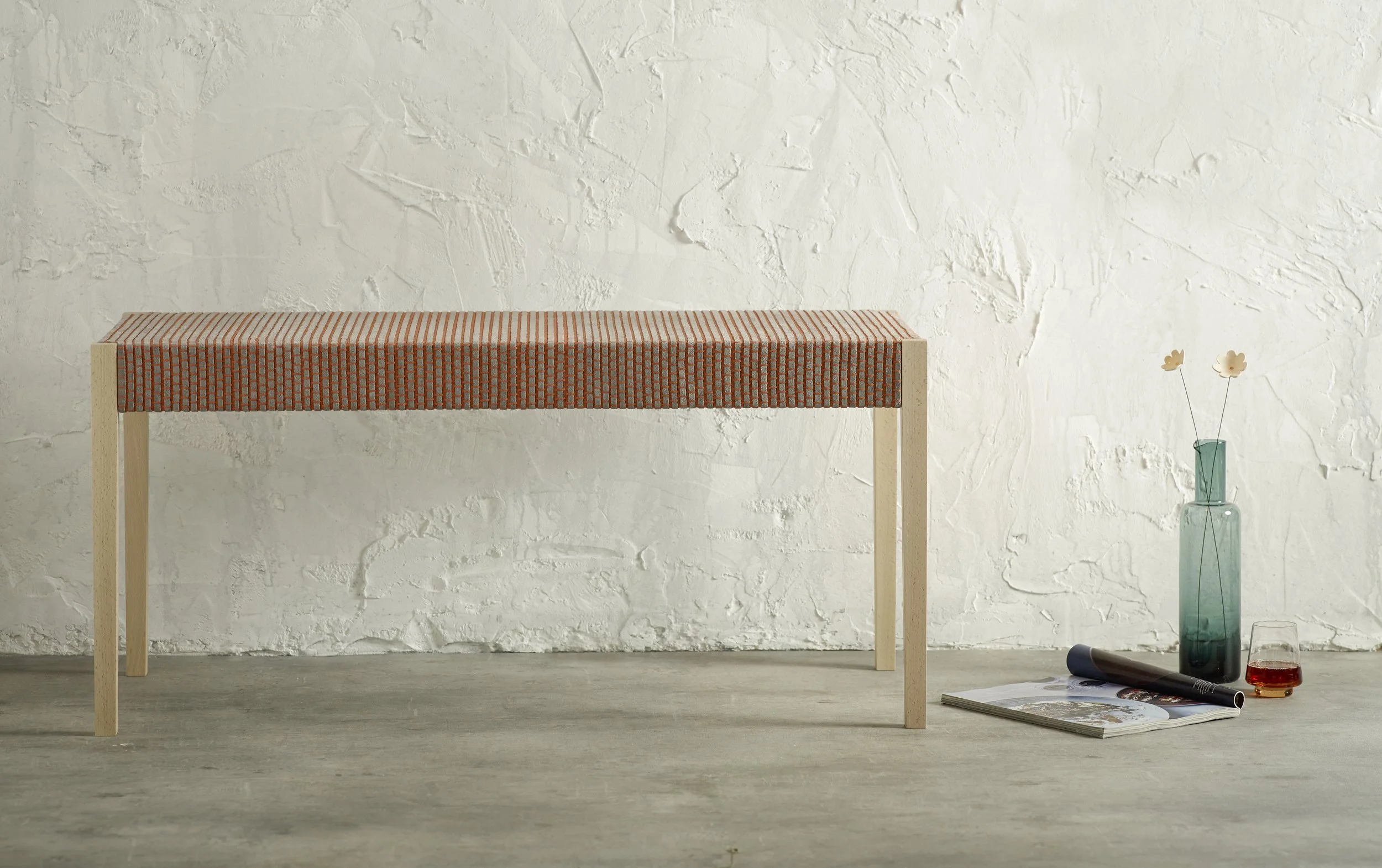 Front view of upholstered bench with orange and grey grid pattern fabric and natural wood legs, beside a vase, magazine, and glass on concrete floor.