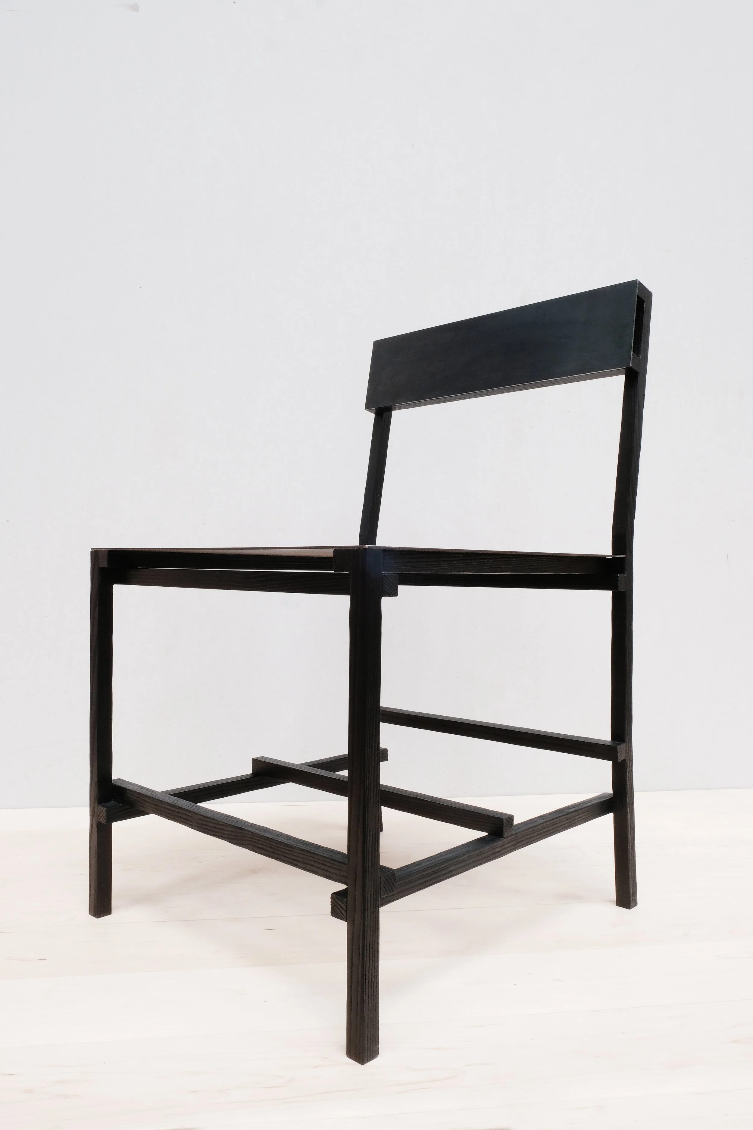A minimalist black wooden chair frame with a wide rectangular backrest, shown at an angle on a light wooden floor.