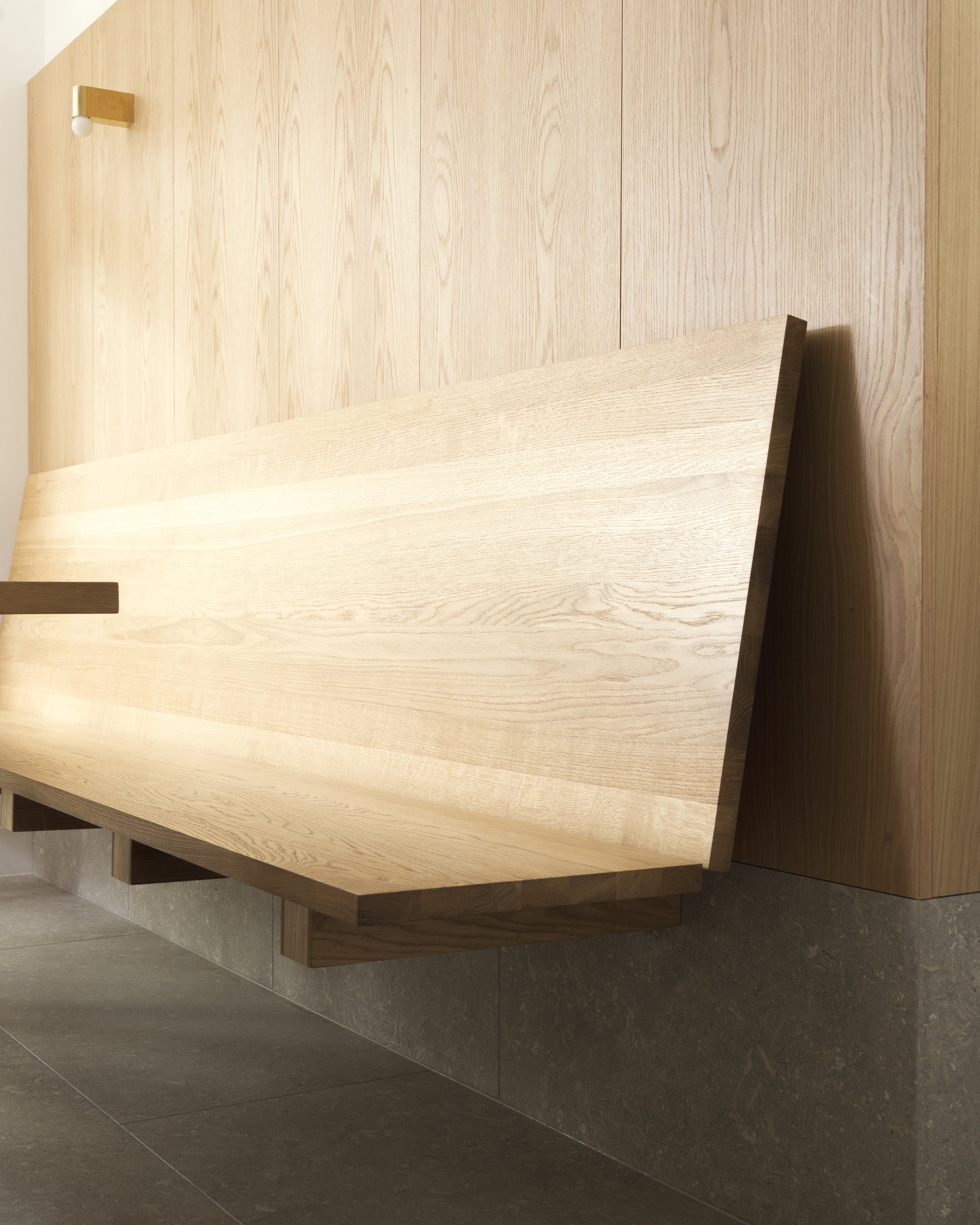 Close-up of cantilevered oak bench seat with sharp chamfered edge mounted on stone-clad wall.