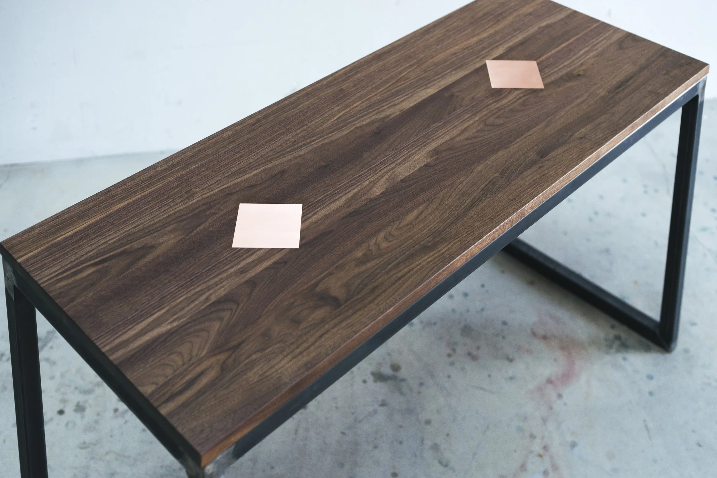 Oblique view of a rectangular bench with dark wooden top and two copper inlays, supported by black metal legs.