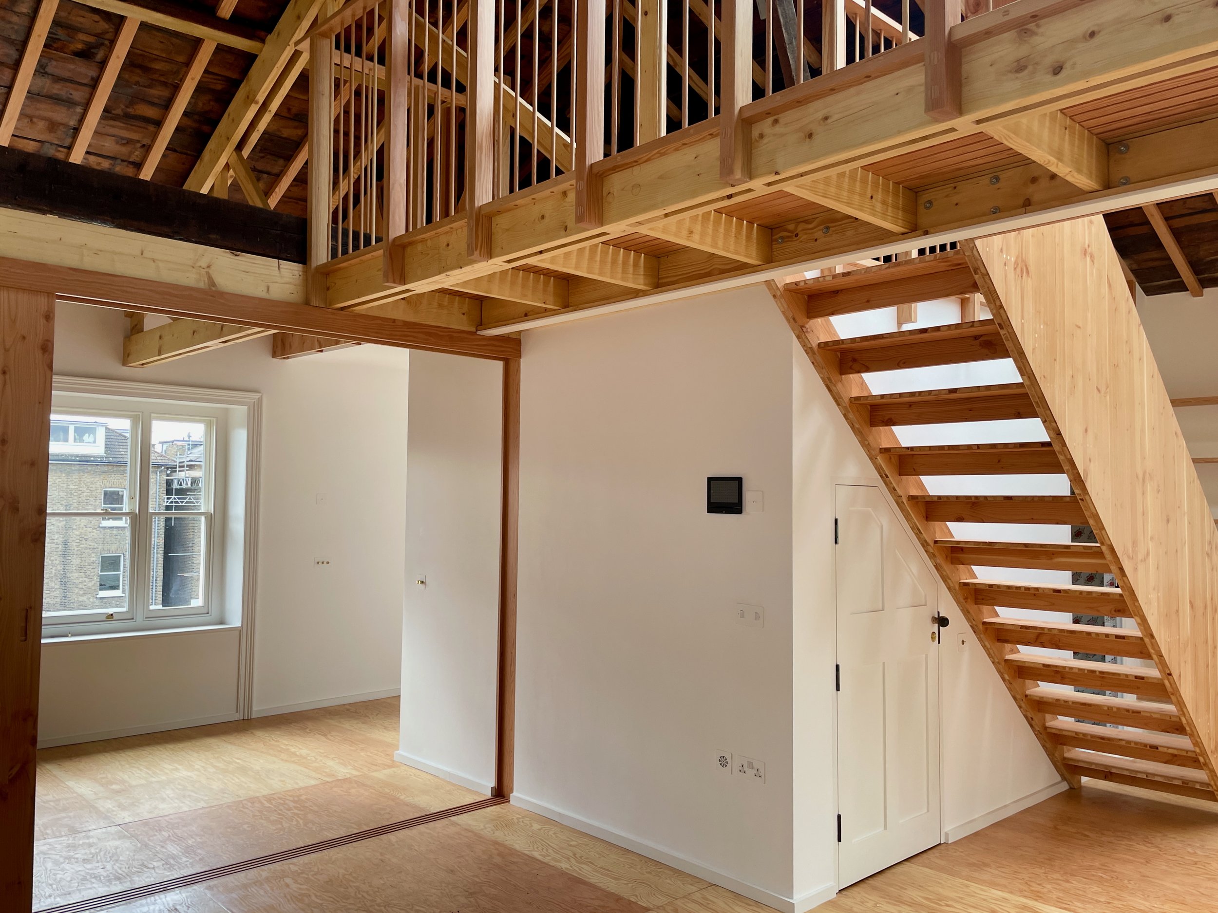 Detail view of a timber mezzanine and staircase structure with natural wood balustrade and a view into a bright adjoining room with sash window.