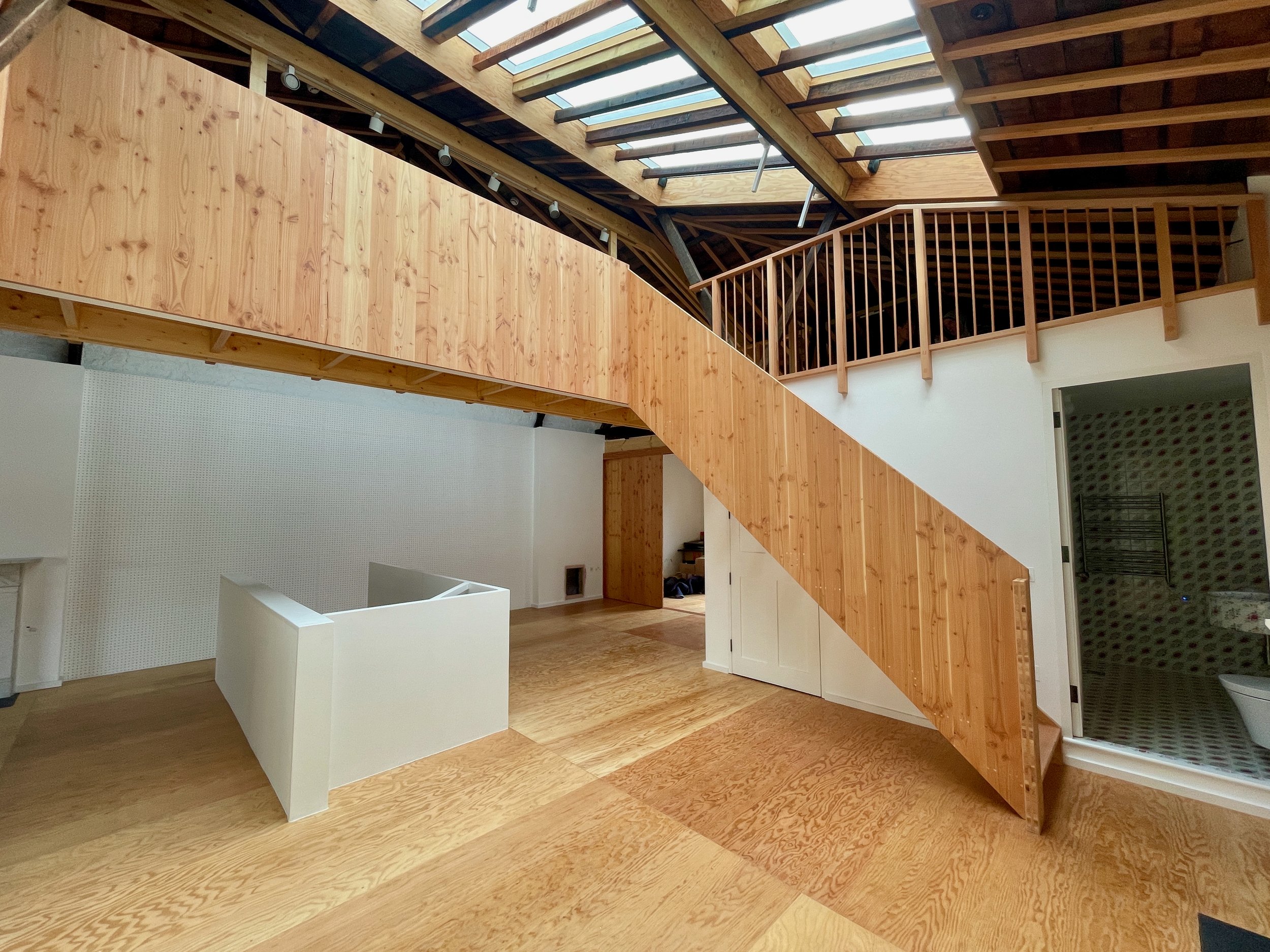 Interior view of a loft-style space with exposed timber roof, Douglas fir staircase and balustrade, and plywood flooring, featuring skylights and white partition walls.