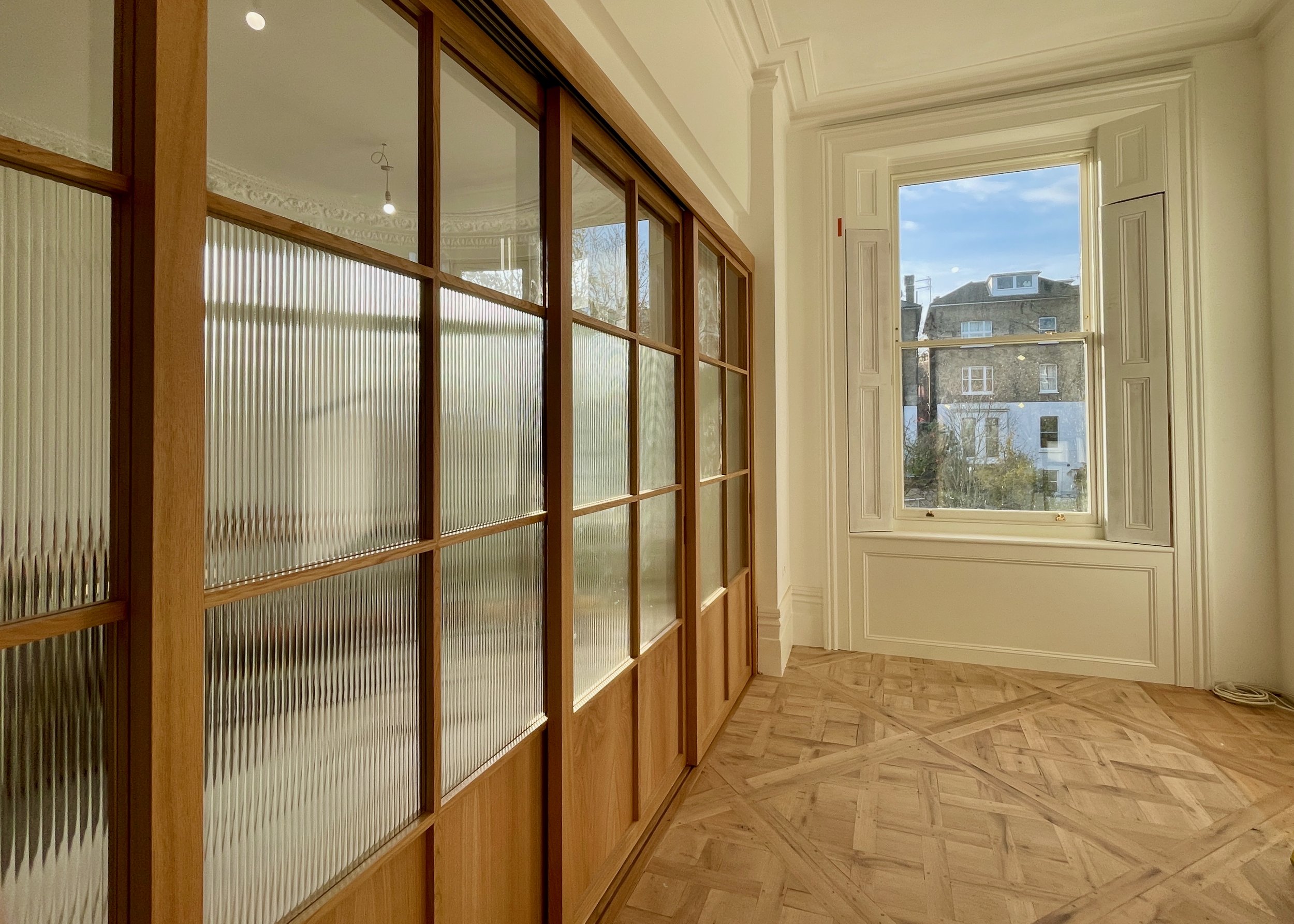 Interior hallway with fluted glass sliding doors framed in wood, parquet flooring, and a large sash window at the end.