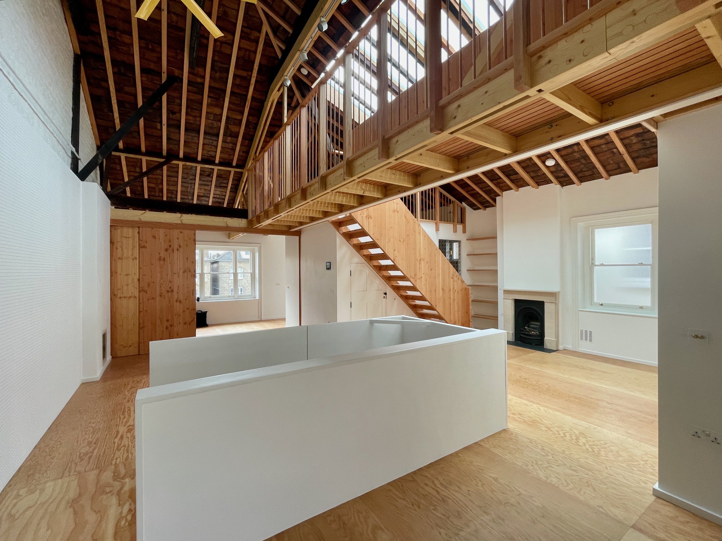 Wide view of an open-plan living space with vaulted ceilings, visible timber beams, a central staircase, and built-in shelving beside a fireplace.