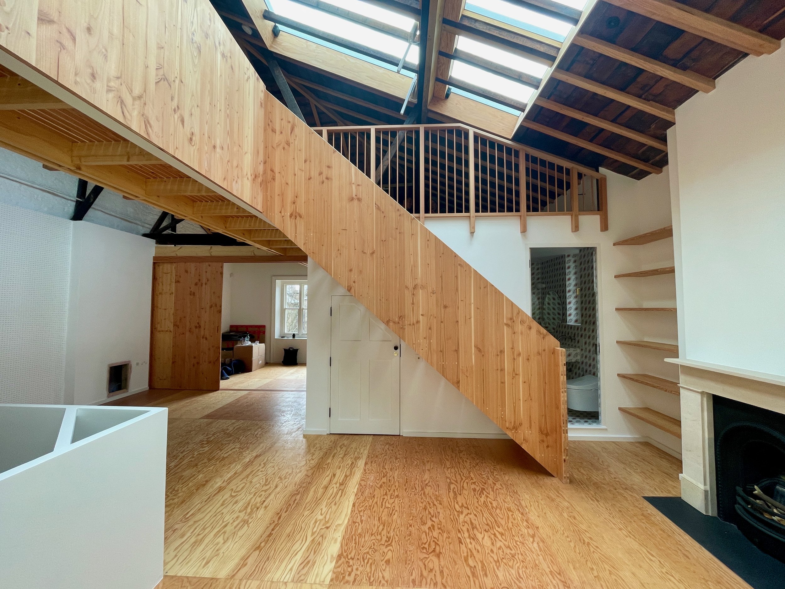 Interior view of a loft-style living space featuring a timber staircase, exposed wooden beams, skylights, and built-in shelving next to a fireplace.