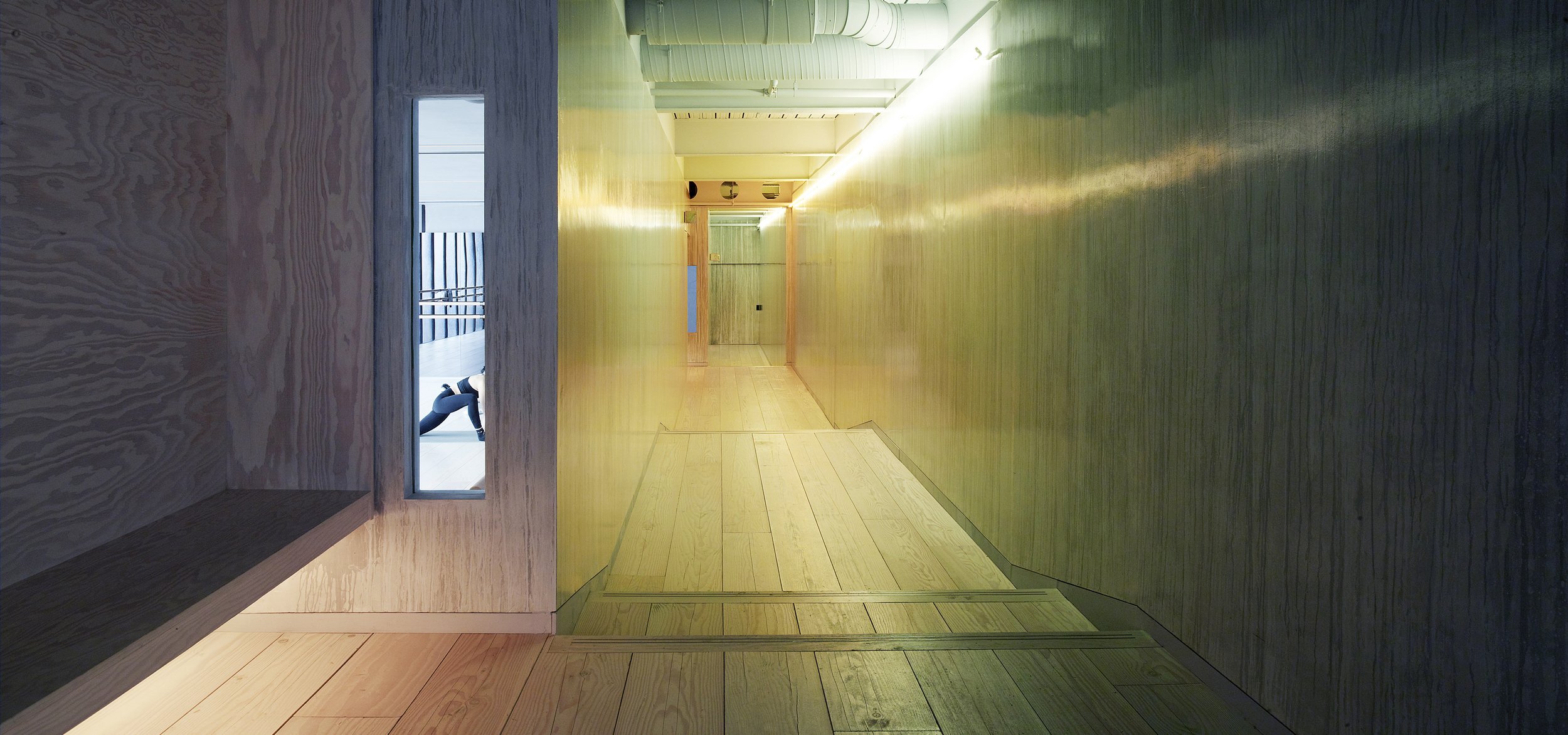 Atmospherically lit wood-lined corridor with built-in bench, stepped floor, and a small window revealing a person stretching in a Pilates studio.