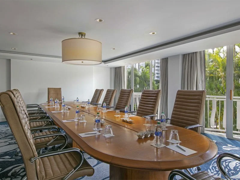 Conference room with a long wooden table, high-back leather chairs, water bottles, glasses, notebooks, and a large window with curtains showing greenery outside.