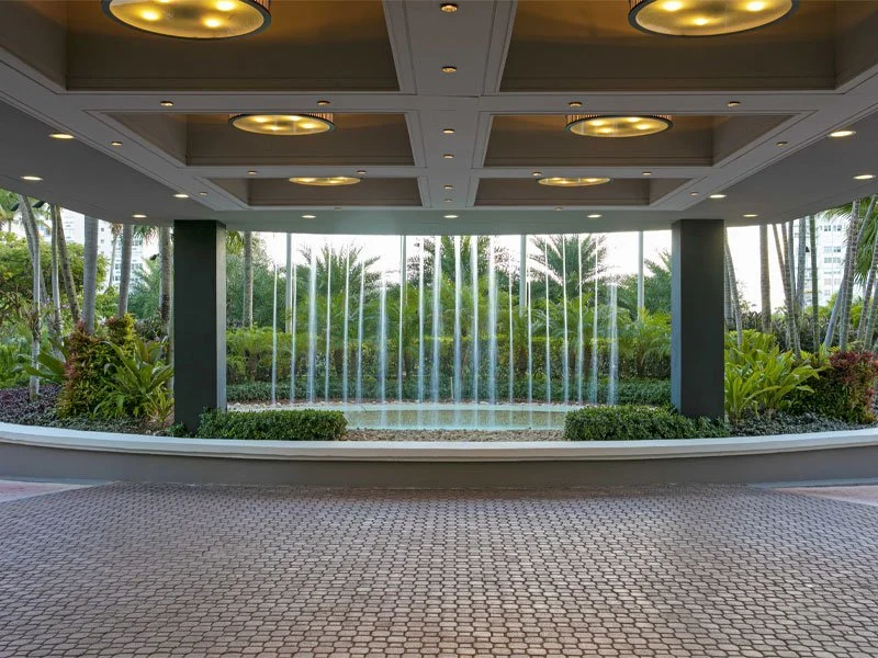Indoor area with a decorative water fountain, lush green plants, and a view of trees and city buildings through large glass windows.