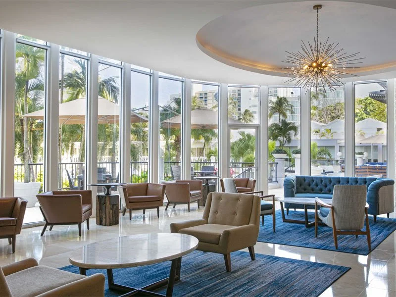 Interior of a modern lounge or lobby with large glass windows, stylish seating including beige, blue, and brown chairs, a round coffee table, hanging light fixture, and an outdoor patio with umbrellas visible through the windows.