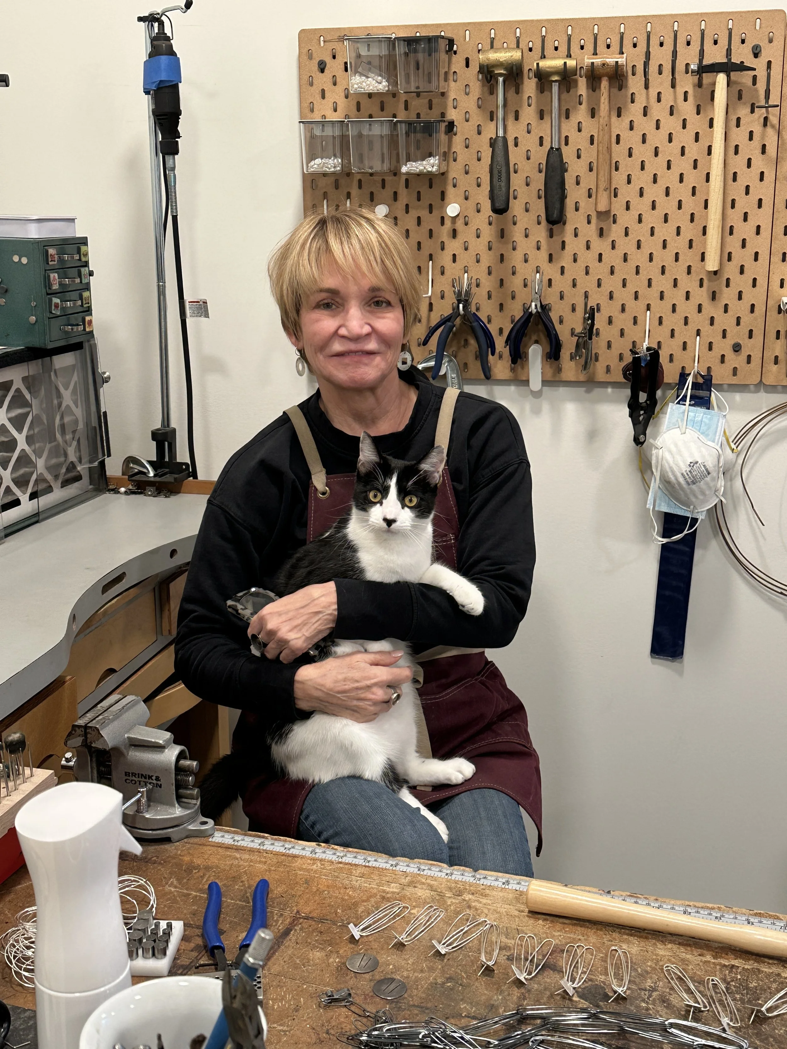 A female metalsmith sitting at a workbench with a black and white cat on her lap, surrounded by jewelry-making tools and wire, with a pegboard on the wall holding hammers, pliers, and boxes with small parts.